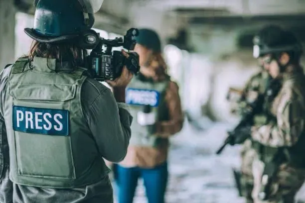 A press crew in body armour filming alongside armed personnel in a hostile environment